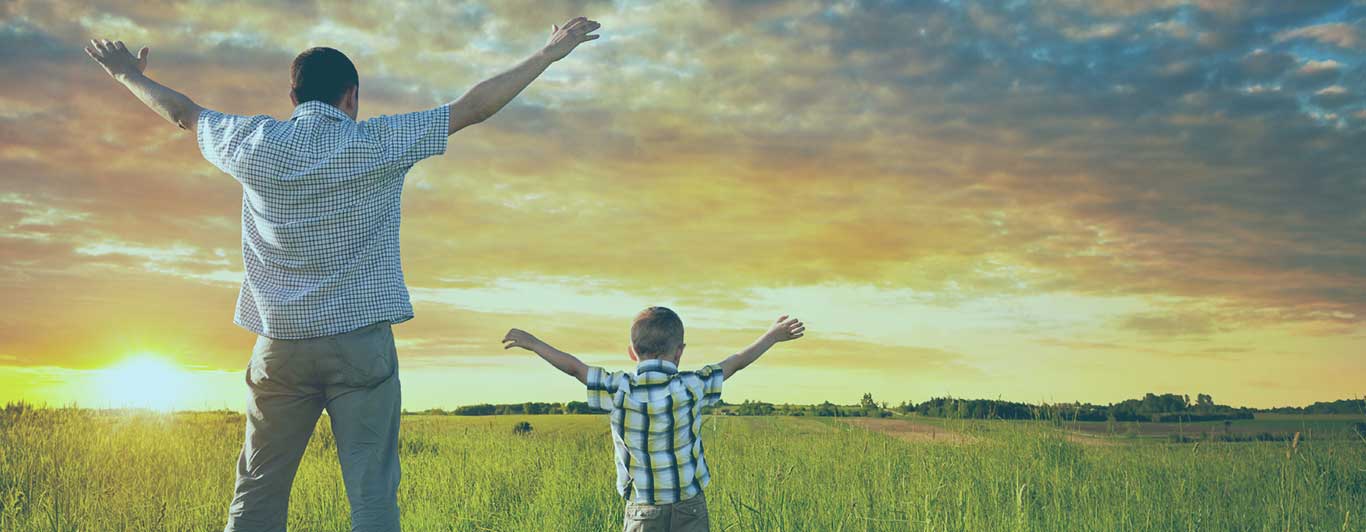 A father and child raising their hands in an open field overlooking the green vegetation.